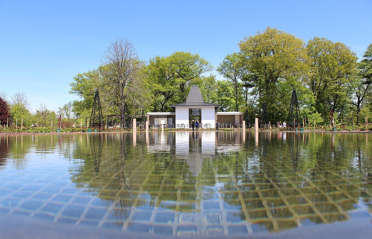 Daytime view of trees and a white building reflected in a pond at Hendrie Park beneath a blue sky
