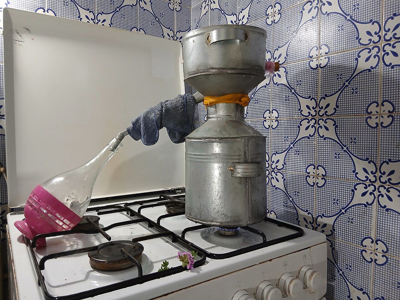 A metal still heated on a gas range in a blue-tiled kitchen, liquid traveling through a tube into a bottle, with a sprig of rose geranium in front